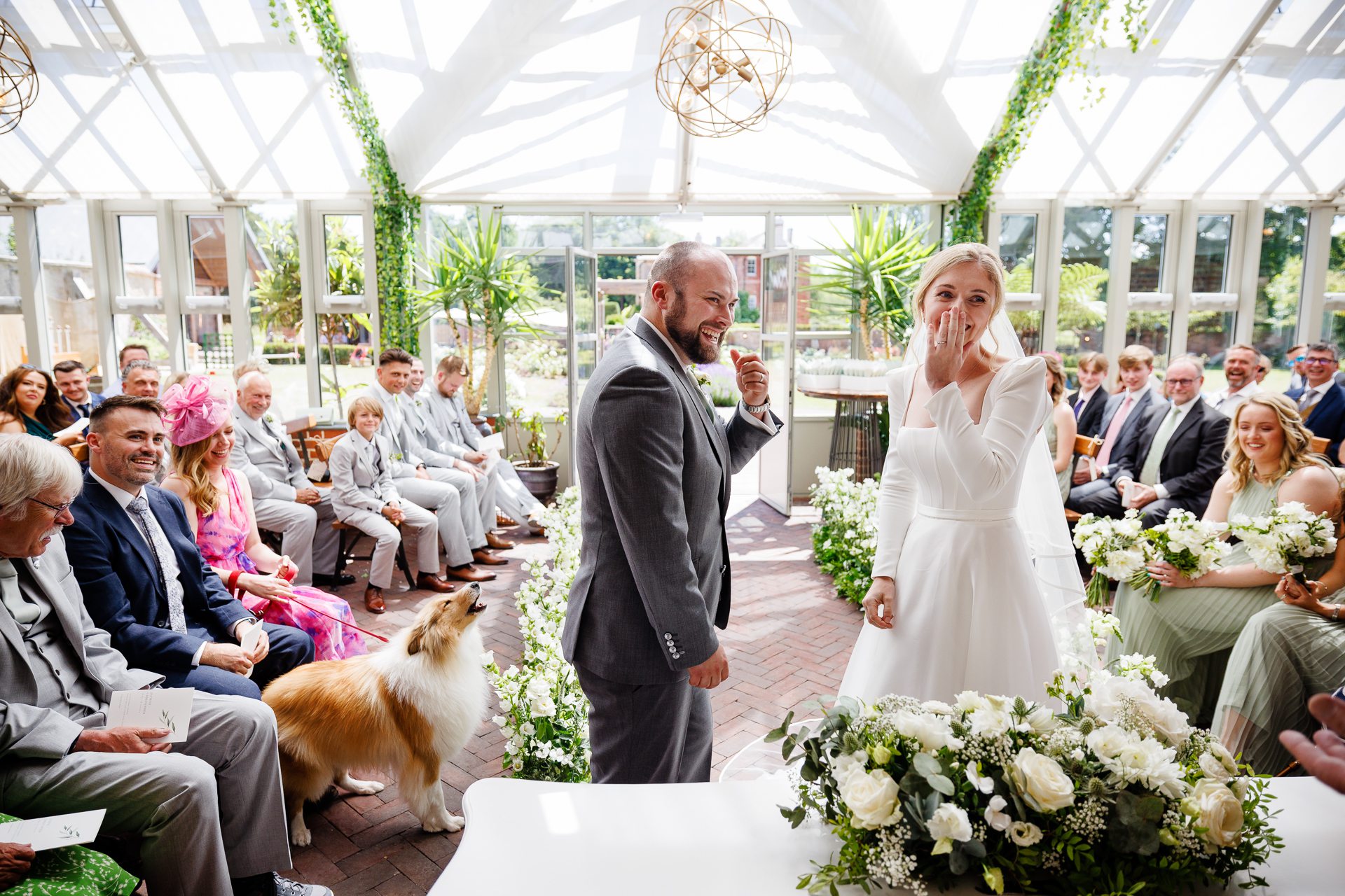 A bride and groom laugh as their dog woofs during their summer syrencot wedding in the glasshouse.